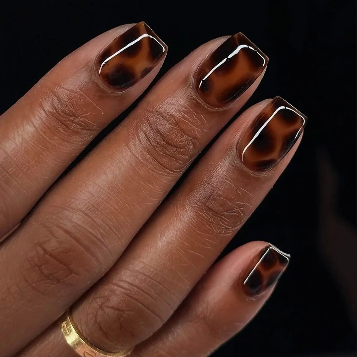Short square press-on nails in a glossy dark brown tortoiseshell pattern on a hand with deep skin tone, with application kit.